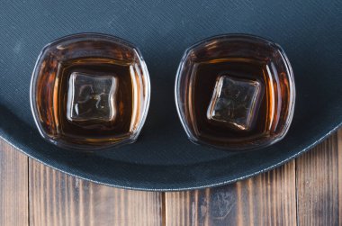 Siyah tepside buzlu viski bardakları ve ahşap masa / Siyah tepside buz küpleri olan iki bardak viski ve kopyalanmış ahşap masa. Üst görünüm.