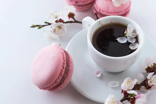 Pink macaroons with a cup of coffee and a branch of white flowers on a white background. French dessert and flowers blooming cherry. Sakura flowers with pink macaroon close up.