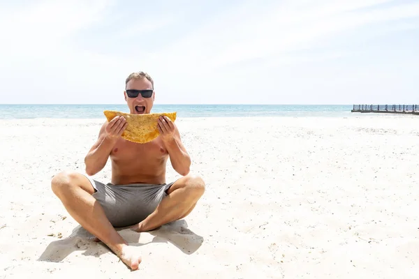 Happy man eating meat pastry Cheburek against sea beach background. Beach food concept