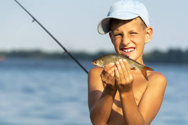 Feliz niño sonriente sosteniendo pescado de agua dulce tomado en las manos contra la caña y el fondo del río — Foto de Stock