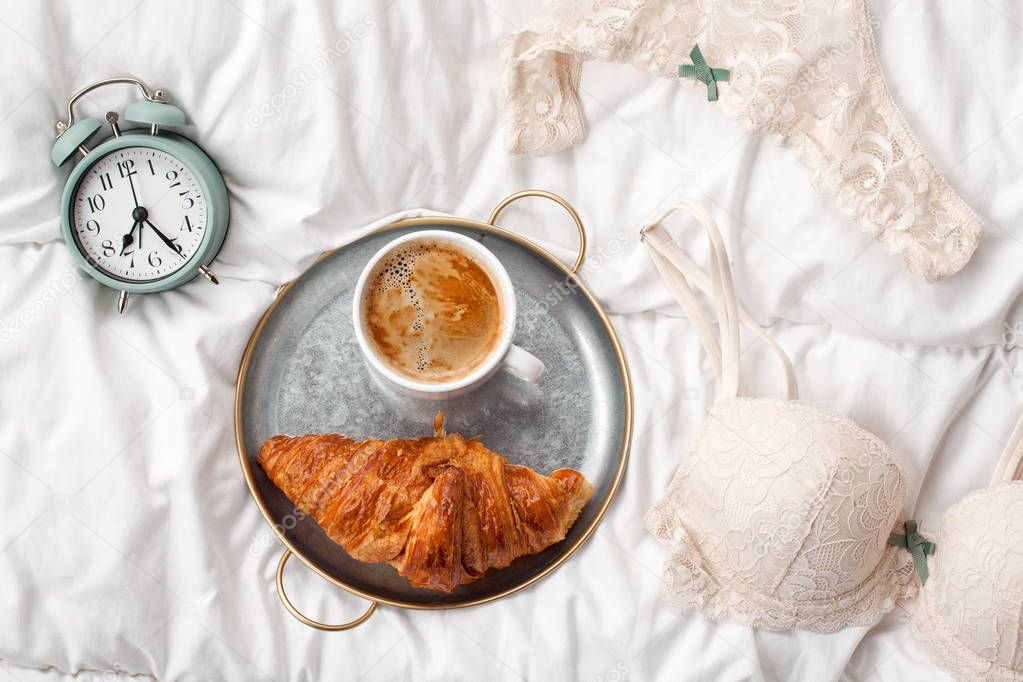 Coffee with croissant, alarm clock and girls underwear in the bed at home . Morning time and breakfast in bed background concept with copy space