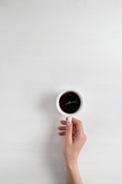 Genç bir kızın eli, açık ahşap bir arka planda bir fincan kahve tutuyor. Sabah kahvesinin içindeki çalar saati çevirin. Günaydın konsept.