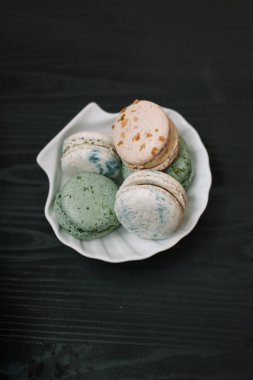 Siyah ahşap arka plandaki beyaz tabakta renkli ev yapımı badem kurabiyeleri duruyor. Desen.