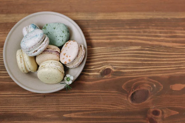 Kahverengi ahşap arka plandaki bej tabakta renkli ev yapımı badem kurabiyeleri var. Desen.