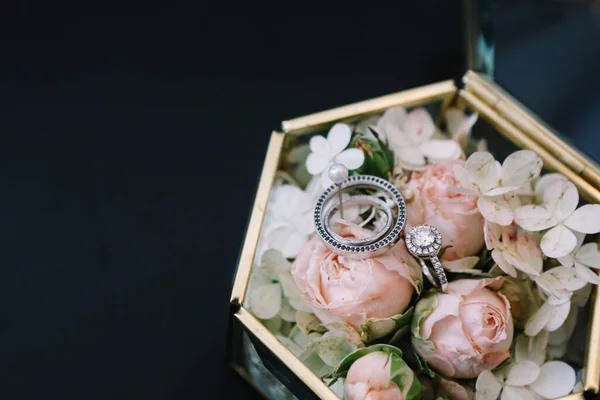 有粉色玫瑰 白色花朵和结婚戒指的老式玻璃盒放在黑色桌子的背景上 — 图库照片