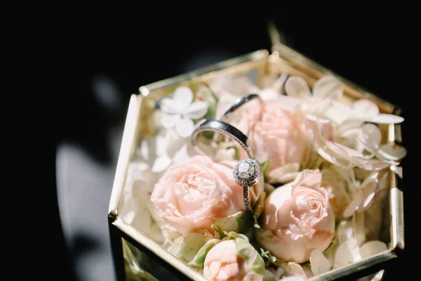 有粉色玫瑰 白色花朵和结婚戒指的老式玻璃盒放在黑色桌子的背景上 — 图库照片