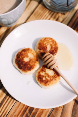 Lezzetli üç süzme peynirli krep. Beyaz tabağın üzerinde bal var. Ahşap masa zemininde çiçekli bir ağacın dalında kahve fincanı var. Peynirli börek..
