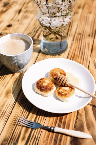 Delicious three cottage cheese pancakes with honey lying on the white plate with the branch of a flowering tree on the wooden table background with the cup of coffee. Curd cheese fritters.