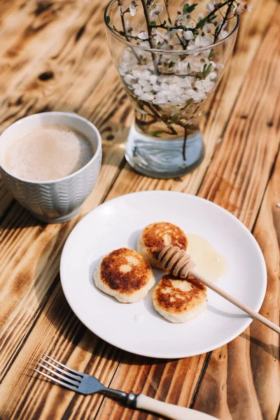 Delicious three cottage cheese pancakes with honey lying on the white plate with the branch of a flowering tree on the wooden table background with the cup of coffee. Curd cheese fritters.