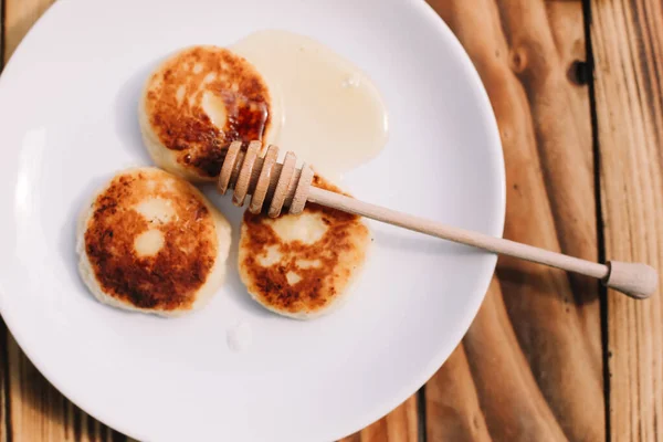 Delicious three cottage cheese pancakes with honey lying on the white plate with the branch of a flowering tree on the wooden table background. Curd cheese fritters.