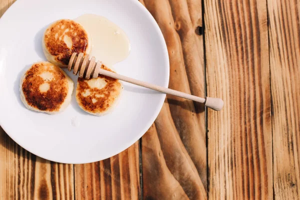 Delicious three cottage cheese pancakes with honey lying on the white plate with the branch of a flowering tree on the wooden table background. Curd cheese fritters.