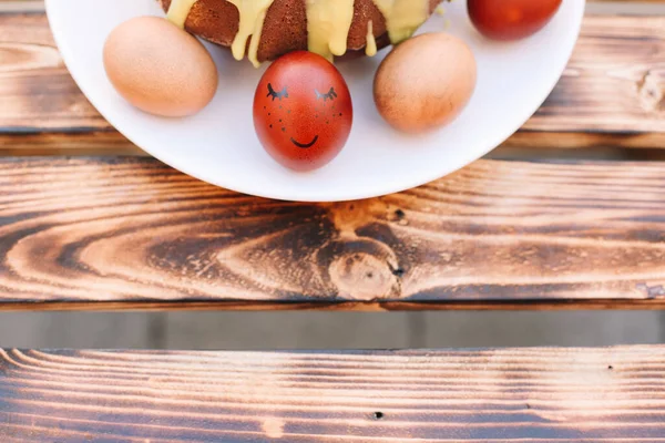 Red easter egg with a smile around eggs and Easter cake in the white plate on the wooden background