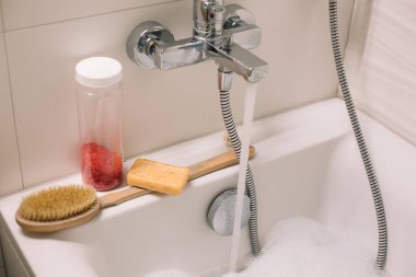 Banyo aksesuarları banyonun üzerinde duruyor. Masaj ve banyo ürünleri, deniz tuzu ve sabun. Organik kozmetik ürünleri. Küvette köpük köpüklü su akıyor..