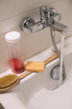 Banyo aksesuarları banyonun üzerinde duruyor. Masaj ve banyo ürünleri, deniz tuzu ve sabun. Organik kozmetik ürünleri. Küvette köpük köpüklü su akıyor..