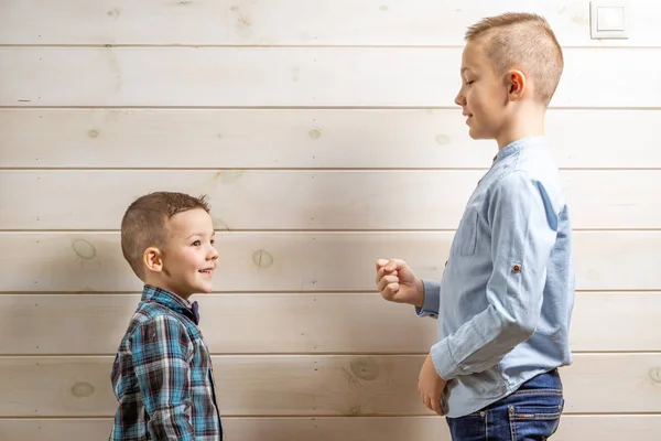 A 4-year-old boy in a blue klepy shirt cries on a light wooden background and his 10-year-old brother stands and plays rock-paper-scissors.
