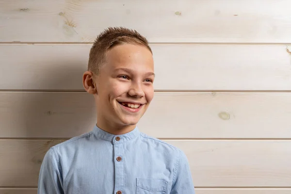 Ένα αγόρι 10 ετών με μπλε πουκάμισο χαμογελά σε ανοιχτό ξύλινο φόντο και κάνει διάφορα σημάδια με τα χέρια του. — Φωτογραφία Αρχείου