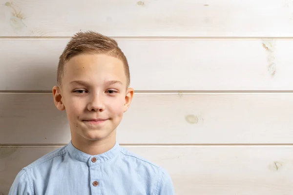Ένα αγόρι 10 ετών με μπλε πουκάμισο χαμογελά σε ανοιχτό ξύλινο φόντο και κάνει διάφορα σημάδια με τα χέρια του. — Φωτογραφία Αρχείου