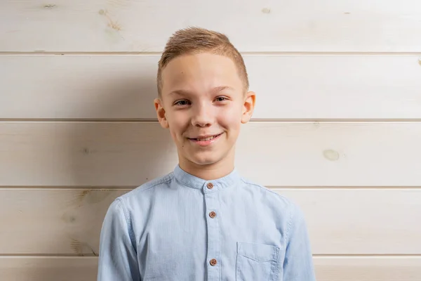 Un garçon de 10 ans dans une chemise bleue sourit sur un fond en bois clair et fait divers signes avec ses mains . — Photo