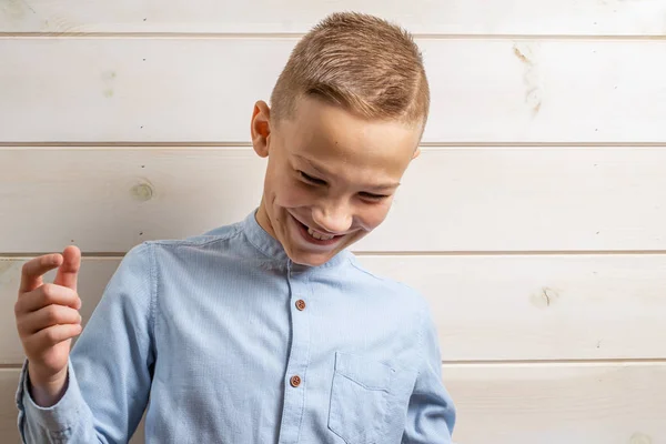 Un garçon de 10 ans dans une chemise bleue sourit sur un fond en bois clair et fait divers signes avec ses mains . — Photo