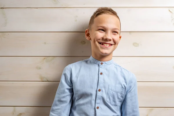 En pojke på 10 år i blå skjorta ler mot en ljus träbakgrund och gör olika skyltar med händerna. — Stockfoto
