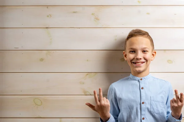 En pojke på 10 år i blå skjorta ler mot en ljus träbakgrund och gör olika skyltar med händerna. — Stockfoto