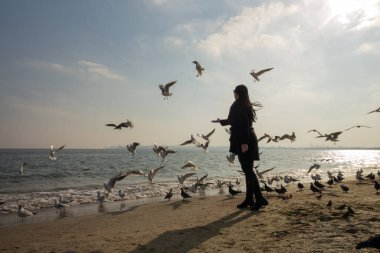 Kumsalda, güneşli bir bahar gününde, siyah ceketli, uzun saçlı esmer bir kadın.