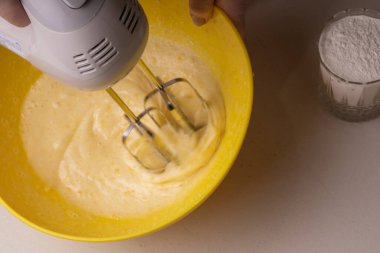 Bir kadın sarı bir kasede turtanın malzemelerini elektrikli mikserle karıştırıyor.