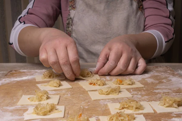 A woman sculpts dumplings and ravioli from squares of dough and cabbage. Plywood cutting board, wooden flour sieve and wooden rolling pin - tools for making dough