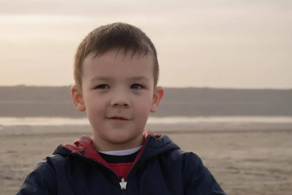 En pojke på 4 år i blå jacka på en tom strand. Barnet ler.. — Stockfoto