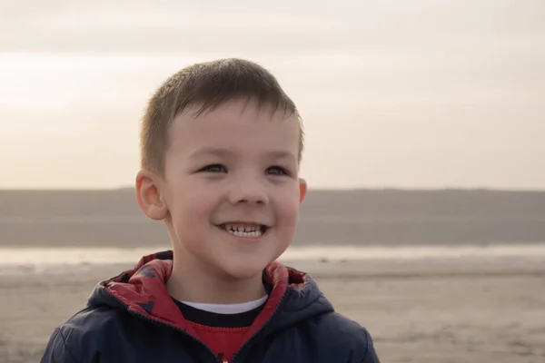 En pojke på 4 år i blå jacka på en tom strand. Visar tänder. — Stockfoto