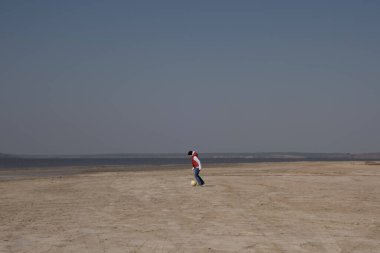 10 yaşında beyaz kazak ve turuncu yelek giyen bir çocuk ıssız bir sahilde futbol oynuyor.