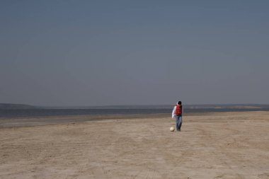 10 yaşında beyaz kazak ve turuncu yelek giyen bir çocuk ıssız bir sahilde futbol oynuyor.