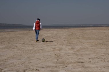 10 yaşında beyaz kazak ve turuncu yelek giyen bir çocuk ıssız bir sahilde futbol oynuyor.