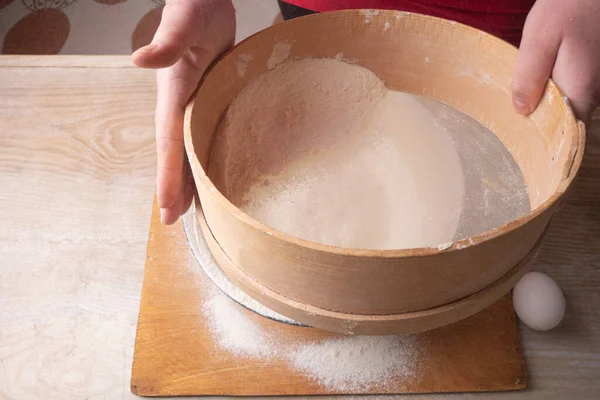 Grand bol, planche à découper, tamis pour tamiser la farine sur la table. Mains féminines tamisant la farine dans la cuisine de la maison . — Photo