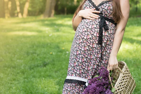 Крупним планом торс молодої вагітної моделі, що стоїть з бузковою квіткою в парку. Майбутня мама очікує, що дитина вийде на вулицю. Концепція здорового материнства . — стокове фото