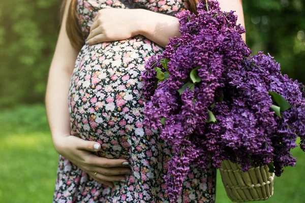 公園でライラックの花と若い妊婦モデル立ちの胴体のクローズ アップ。今後のママ期待して赤ちゃんが屋外で歩きます。健康的な出産の概念. — ストック写真