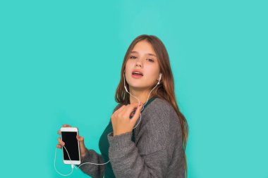 Moda oldukça tatlı, kaygısız genç kız kulaklıkla müzik dinliyor ve mavi arka planda akıllı telefonla şarkı söylüyor.