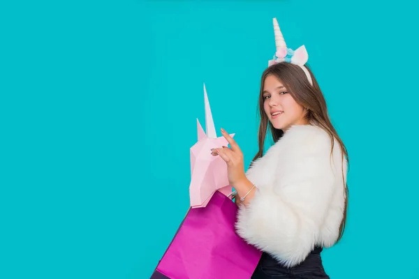 Portrait of a smiling pretty girl with shopping bags over blue background. Black friday concept. Kawaii teenage girl with unicorn horn buying a paper pink unicorn. — ストック写真