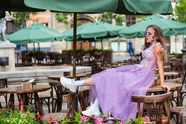 Retrato do jovem gil vestindo vestido e óculos de sol violeta sentado na mesa de cafés. tatuagens nos braços e na cara. Fecha. Caminhada. Atributos da subcultura . — Fotografia de Stock