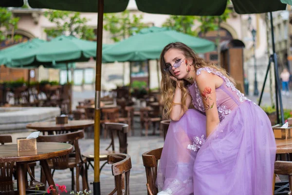 Retrato do jovem gil vestindo vestido e óculos de sol violeta sentado na mesa de cafés. tatuagens nos braços e na cara. Fecha. Caminhada. Atributos da subcultura . — Fotografia de Stock