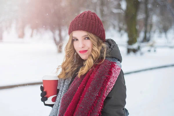 Belle adolescente mignonne et souriante avec maquillage naturel et cheveux bouclés légers et chapeau rouge tient une tasse avec du café chaud ou du thé. Saison d'hiver. Noël, Nouvel An, concept vacances d'hiver . — Photo