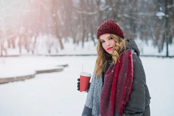 Belle adolescente mignonne et souriante avec maquillage naturel et cheveux bouclés légers et chapeau rouge tient une tasse avec du café chaud ou du thé. Saison d'hiver. Noël, Nouvel An, concept vacances d'hiver . — Photo