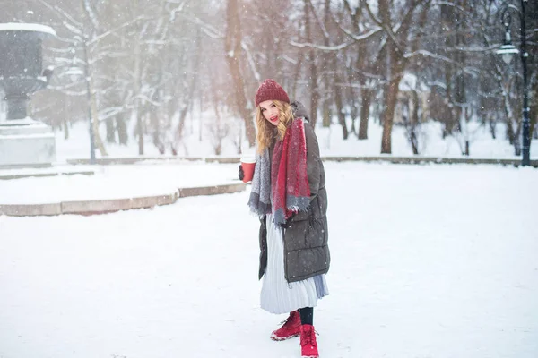 漂亮的笑着可爱的少女，有着自然的妆容和浅色卷曲的头发，戴着红帽子，手里拿着一杯热咖啡或茶。 冬季。 圣诞节、新年、寒假概念. — 图库照片