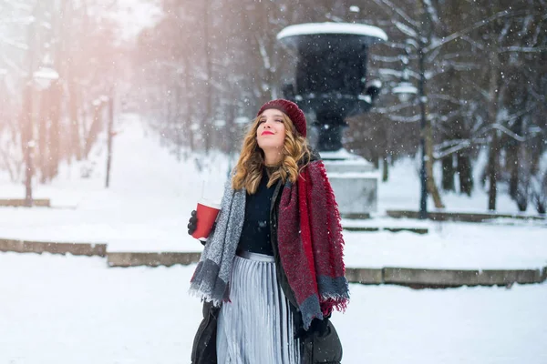 Linda menina adolescente sorridente bonito com maquiagem natural e cabelo encaracolado claro e chapéu vermelho mantém uma xícara com café quente ou chá. Temporada de inverno. Natal, Ano Novo, conceito de férias de inverno . — Fotografia de Stock