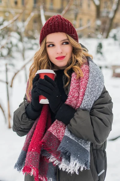 Gyönyörű mosolygós aranyos tizenéves lány természetes make up és könnyű göndör haj és piros kalap tart egy csésze forró kávé vagy tea. Téli szezon. Karácsony, újév, téli ünnepek koncepciója. — Stock Fotó