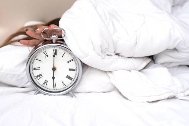 Hand under blanket reaching out for alarm clock, shallow depth of field focus on foreground. Wake up of an asleep girl stopping alarm clock on the bed in the morning