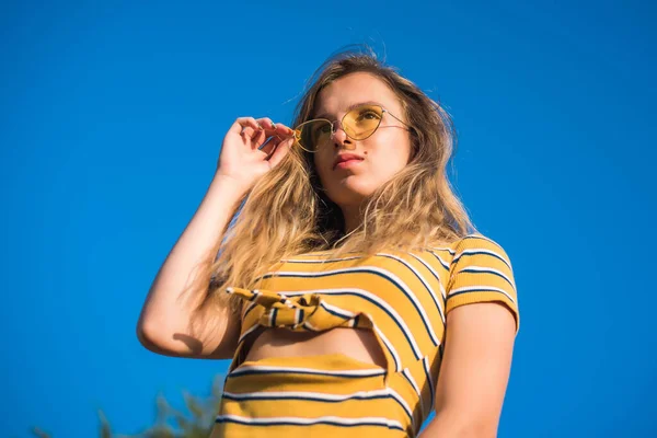 Adolescente feliz menina atraente olha para a câmera na praia ensolarada sobre o céu azul usando óculos de sol amarelos . — Fotografia de Stock
