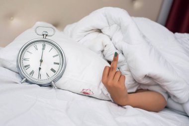 Hand under blanket showig middle finger, offensive gesture. Fuck you concept. for alarm clock, Photo of young couple sleeping in bed while woman stretching hand to alarm clock to turn it of.