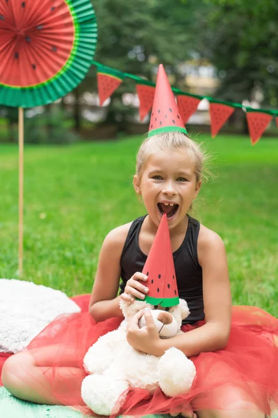 Festa dell'anguria, picnic per bambini nel parco. il giorno dell'anguria. Carino piccola ragazza con cappello e con orso giocattolo con cappello di anguria — Foto Stock