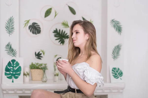 Mujer joven bebiendo té sentado en el hermoso interior con flores verdes en el fondo —  Fotos de Stock